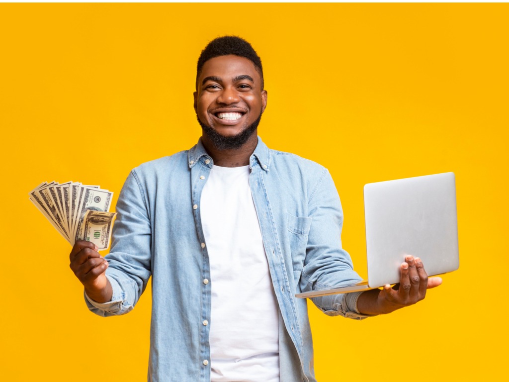 young African American man smiling with money in one hand and laptop in the other hand - How to Make Money Online